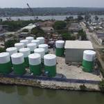 Aerial view of twelve large green and white FRP storage tanks at an industrial facility near a waterfront, surrounded by trees and buildings.