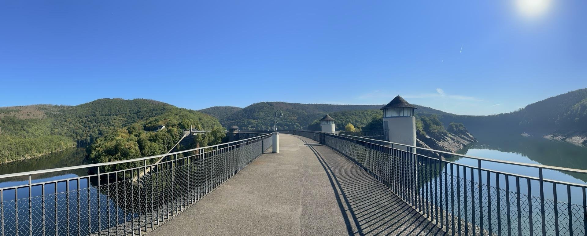 This image shows a scenic view from a dam. The walkway in the image is fenced and leads towards a structure, possibly a control tower or gateway, at the end of the dam. The surrounding area is lush with forested hills, and the reservoir created by the dam is visible on the right side of the image, reflecting the clear sky. The day is sunny and the lighting suggests it might be morning or late afternoon. This setting is likely used both for its functional purpose in water management and as a picturesque spot for visitors to enjoy the view.