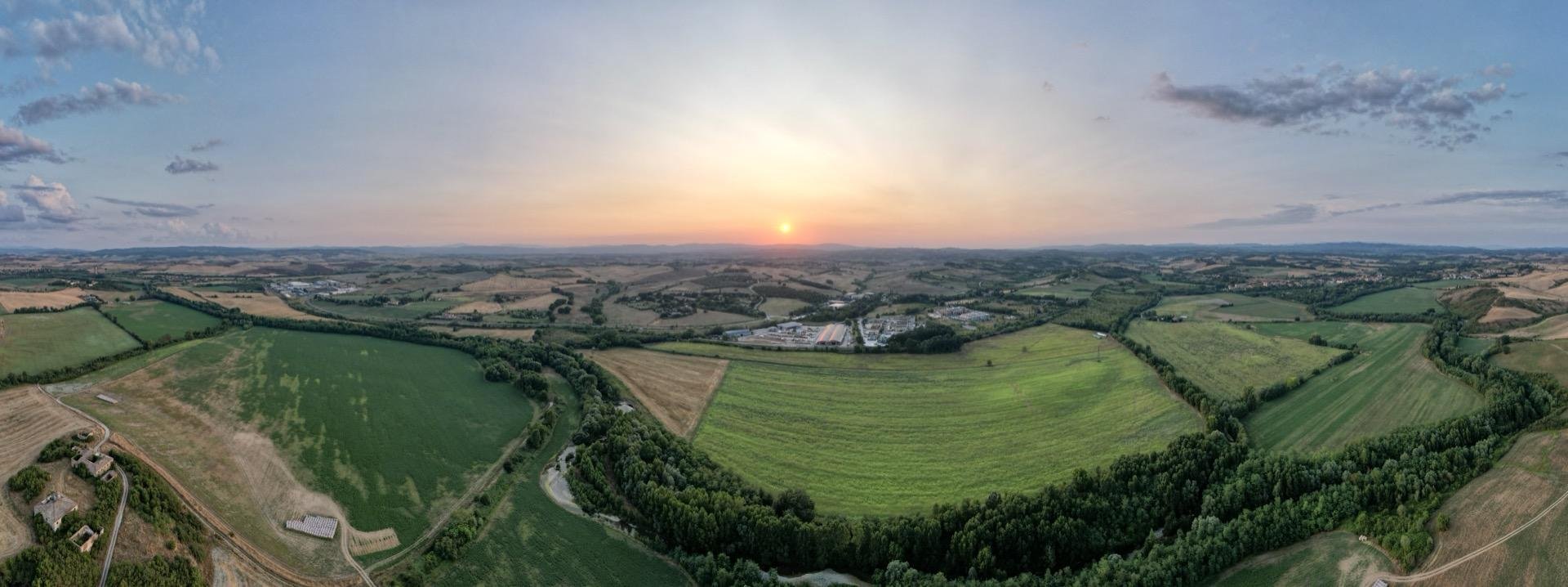 This image is a panoramic aerial view of a rural landscape at sunset. The area features rolling fields with various shades of green, separated by hedgerows and small woodlands. There's a road visible on the left side winding through some of the fields, and a building complex can be seen in the bottom center-right of the image. The horizon is dominated by a clear sky with the sun setting in the center, casting a warm glow and long shadows over the landscape, suggesting a tranquil and spacious rural setting.
