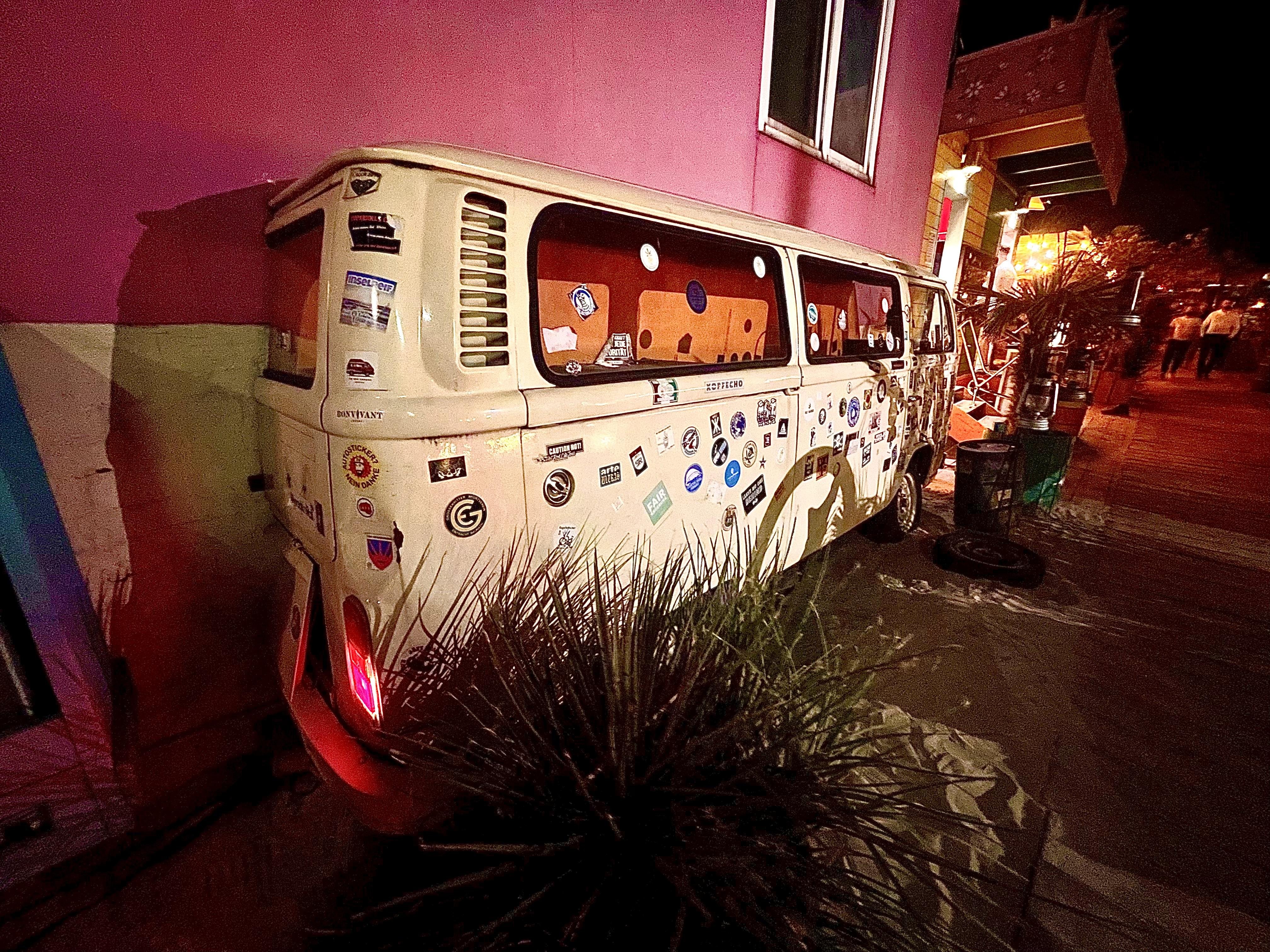 A vintage white Volkswagen bus decorated with numerous stickers parked on a street at night. The van is angled towards a vibrant pink and green wall on its left. A small plant is visible in front, and ambient street lighting casts a dim glow over the scene, with some people and buildings faintly visible in the background.