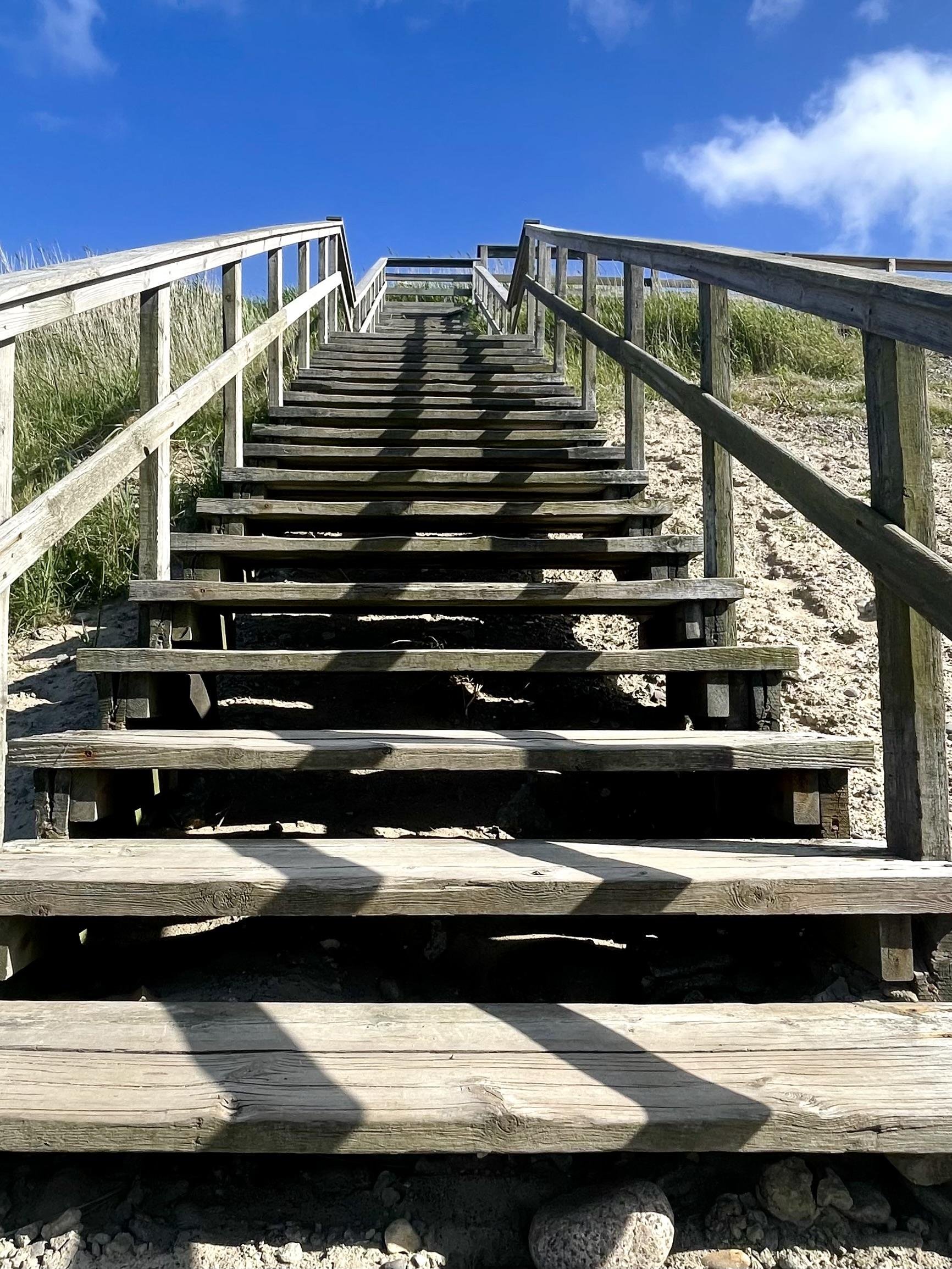 The image shows an upward view of a wooden staircase leading up a sandy slope, likely a dune, toward a bright blue sky with a few clouds. The stairs have handrails on both sides, and you can see grassy vegetation peeking in from the sides, typical of beach or dune flora. The sunlight casts contrasting shadows on the steps, which enhances the texture and dimension of the weathered wood.