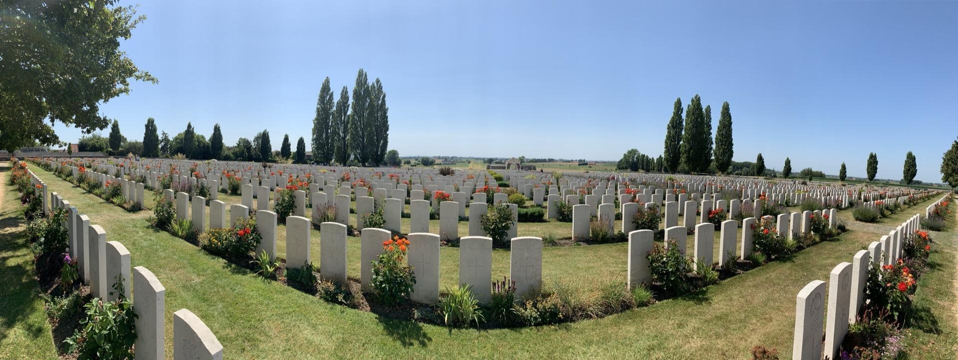 Panoramic view of a war cemetery with rows of uniform white headstones neatly arranged on manicured grass, interspersed with vibrant flower beds under a clear sky. Tall trees line the background, enhancing the serene and orderly landscape.