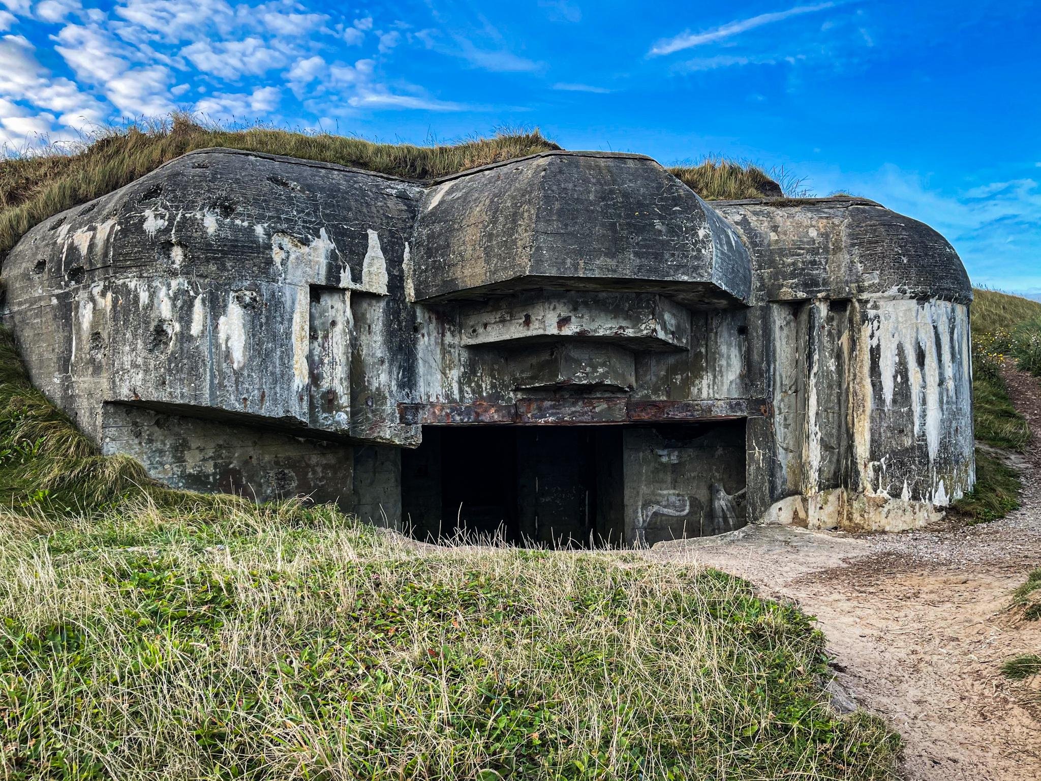 The image features a large, concrete military bunker with a somewhat dome-like structure and reinforced gun emplacements. It is likely from the World War II era, given the style of construction and purpose. The bunker shows signs of aging and weathering, including peeling paint and areas of exposed concrete. The surrounding area appears to be an overgrown grassy field, suggesting it may be located in a coastal or rural setting. Such structures were typically used for defense, equipped to house weapons and soldiers, providing shelter and a strategic vantage point against enemy attacks.