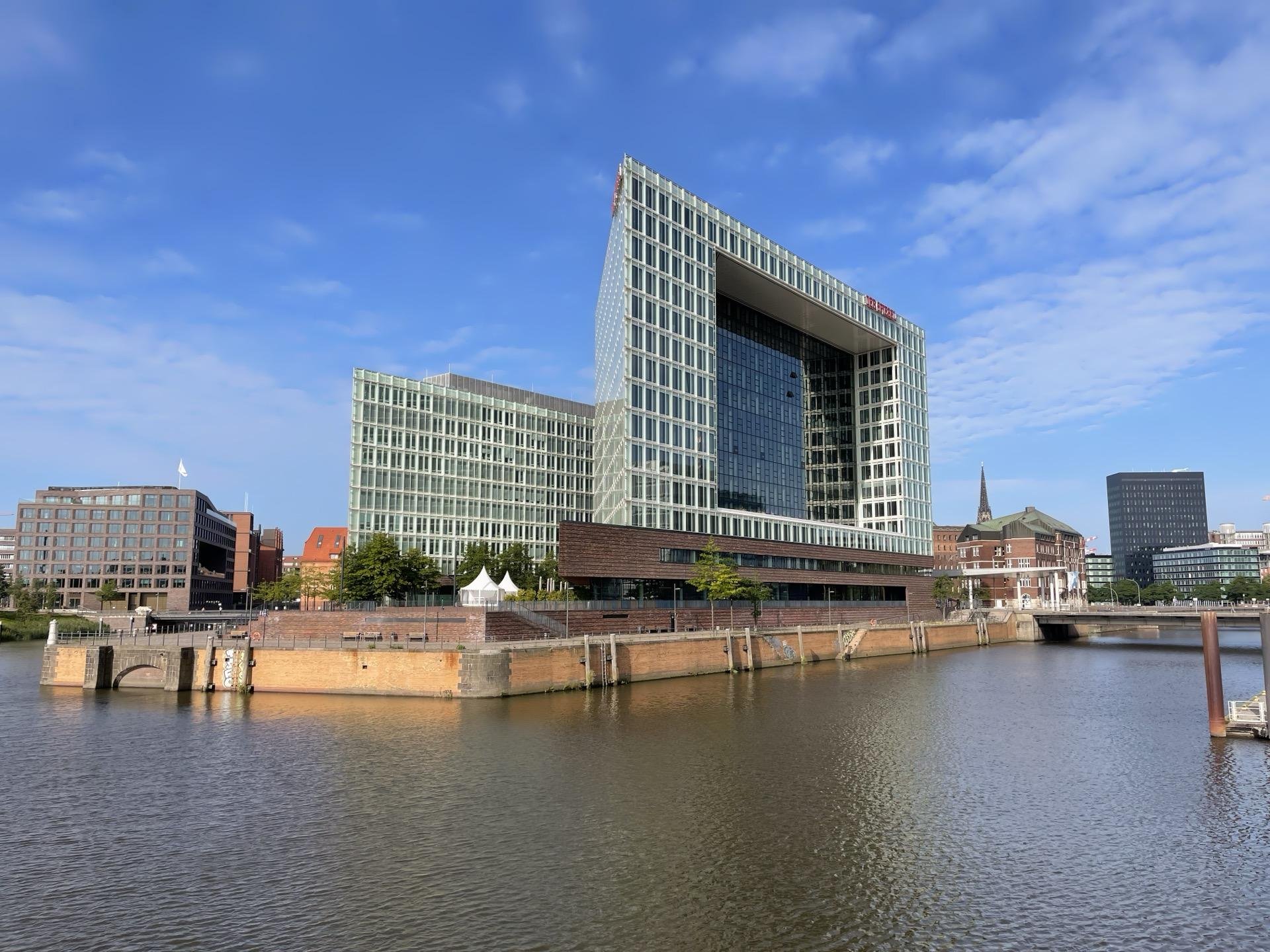 Modern asymmetrical building with expansive glass facade and sharp geometric lines dominates the foreground beside a calm body of water, flanked by other less prominent office buildings under a clear blue sky. An old brick-lined pier extends into the water, contrasting with the contemporary architecture.