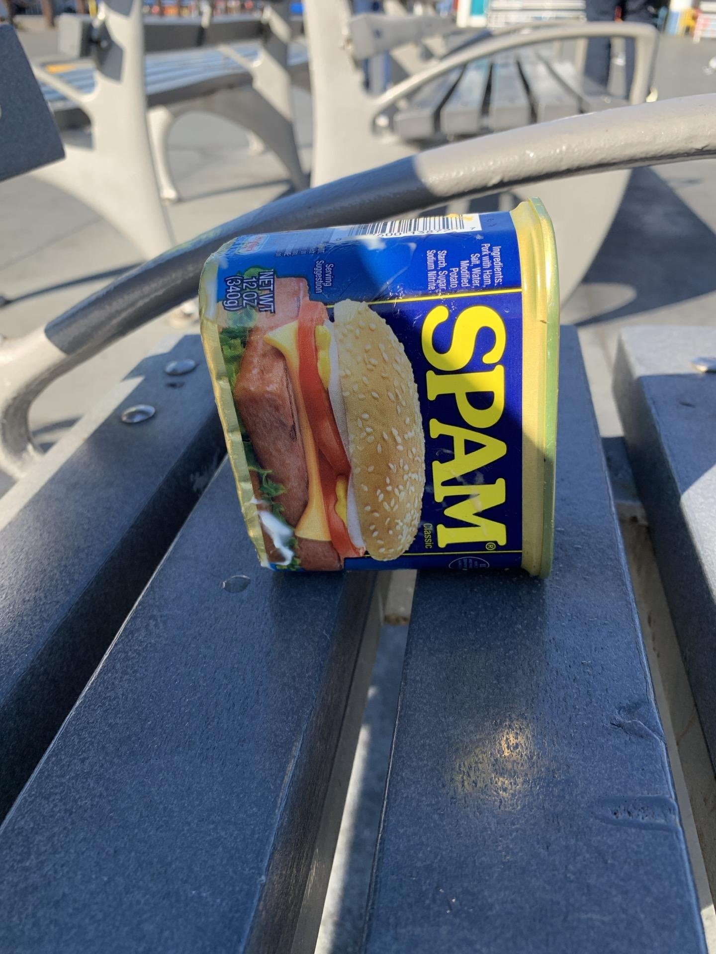 A SPAM canned meat product with a peeling label sits balanced on a dark grey metal bench. The label displays an image of a sandwich. The bench is located in an outdoor setting with exercise equipment in the blurry background. The can is slightly crushed on the side facing the viewer.