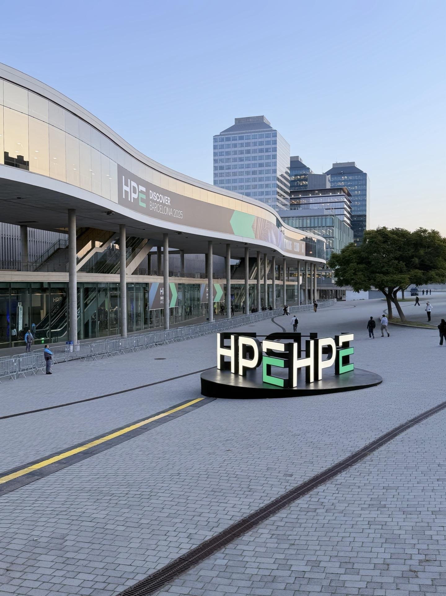 Exterior of a modern conference center with prominent signage reading "HPE Discover Barcelona 2025." A curved building facade sits in the background, adorned with large glass panels and supported by stilts. In the foreground, a circular installation features illuminated "HPE" logos on a black base. Paved public square with several pedestrians, surrounded by tall urban buildings under clear skies.