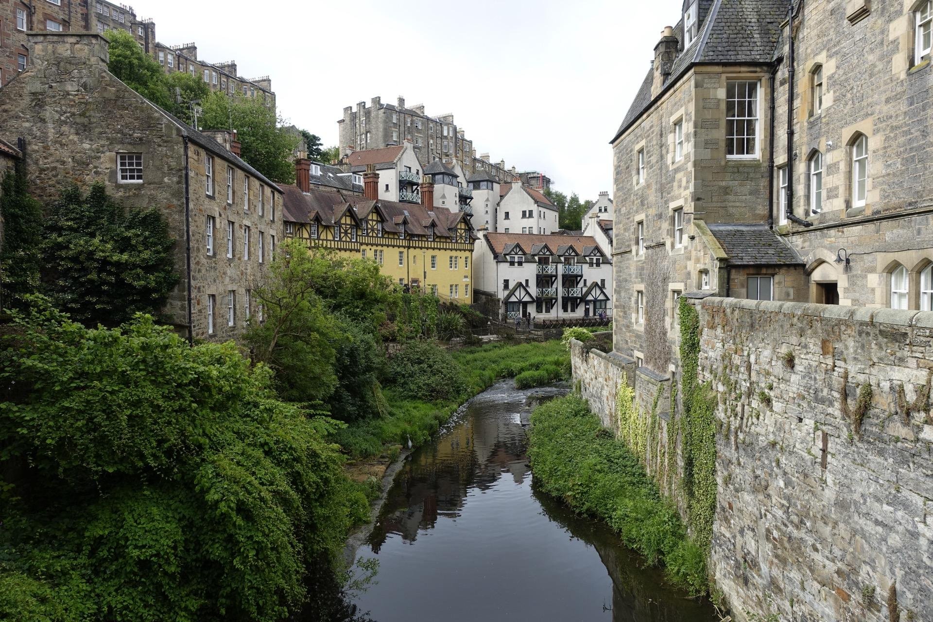 Old stone buildings with varied architectural styles line a narrow river flanked by lush greenery. A quaint footpath runs along the riverbank, with a high stone wall on one side. The structures show a mix of medieval, traditional timber-framed, and Georgian influences, with predominant hues of beige, yellow, and dark timber. The scene is serene and picturesque, set against a soft, overcast sky.
