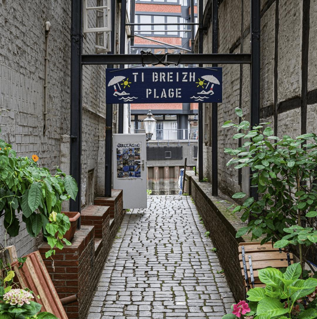 Narrow cobblestone alley flanked by brick and stone walls in an urban setting. A blue sign reading "Ti Breizh Plage" spans across the alley, adorned with white illustrations of beach umbrellas. To the left, green and flowering plants border the path, enhancing the quaint, picturesque atmosphere. The alley leads towards a brighter, more open area with modern buildings in the background.