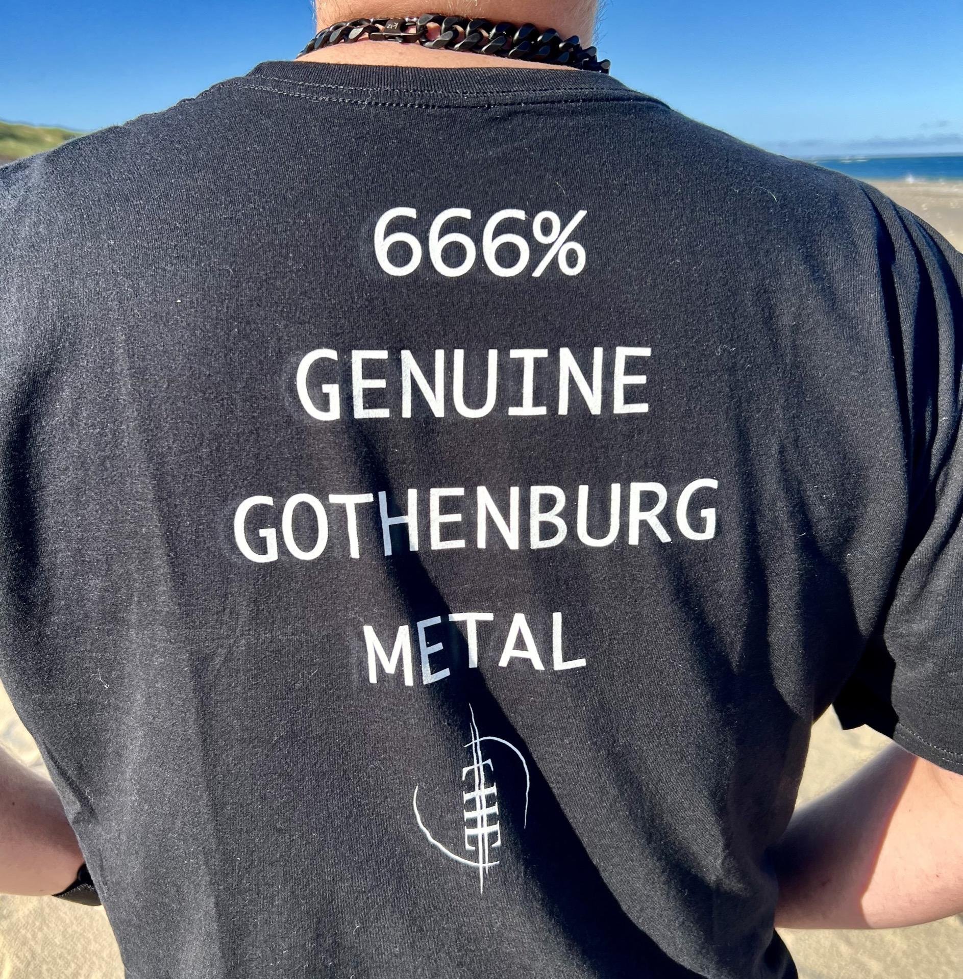 Rear view of a person wearing a black t-shirt with white text reading "666% GENUINE GOTHENBURG METAL" and a small white graphic of a spine below the text. The person is outside, with a clear sky and blurred beach scenery visible in the background. They are wearing a spiked bracelet on one wrist.