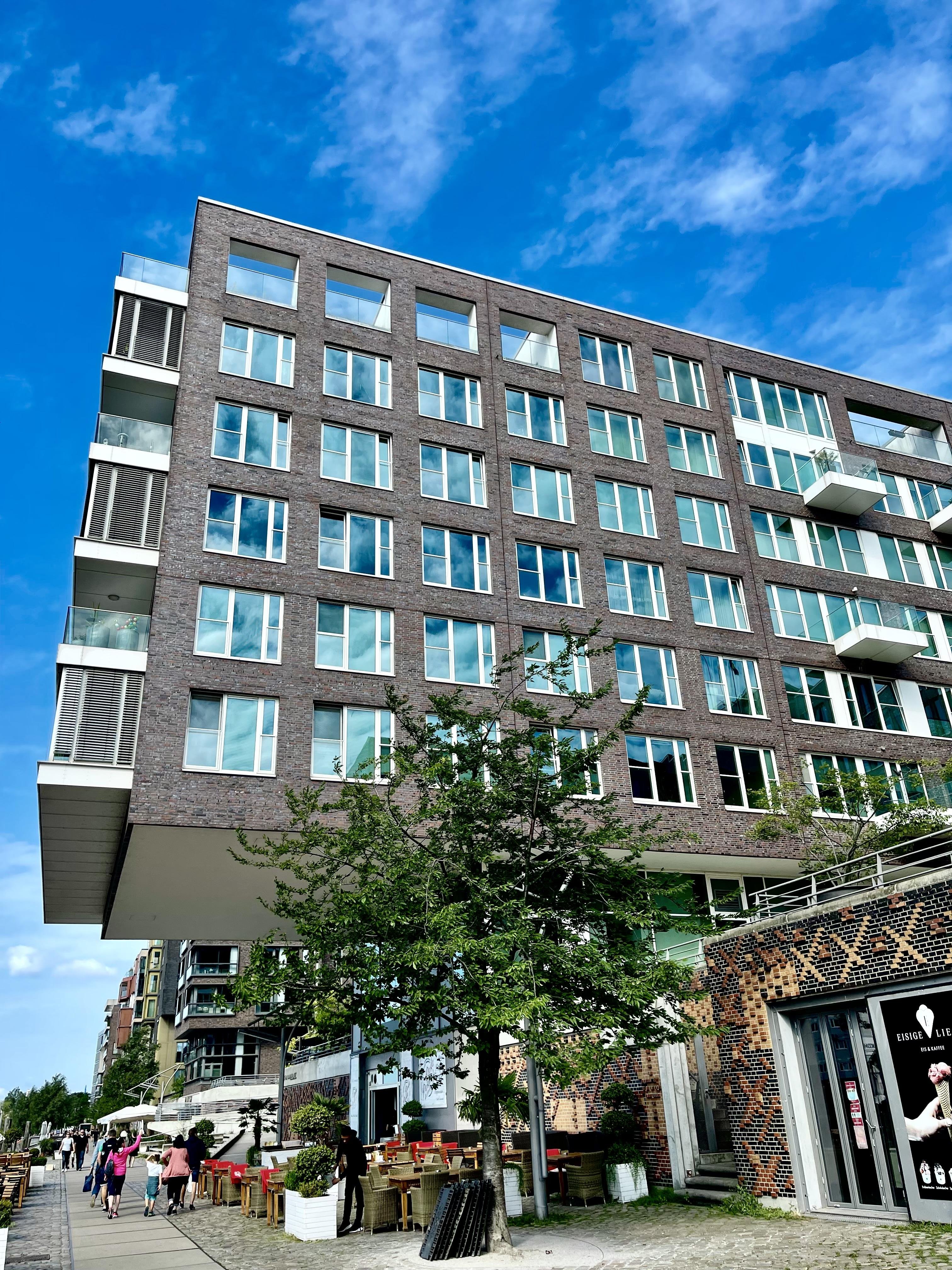 Modern multi-story building featuring a dark brick facade with rows of windows and balconies, under a blue sky with scattered clouds. In the foreground, a lively street scene includes people walking and sitting at outdoor cafe tables lined along the sidewalk, shaded by trees.