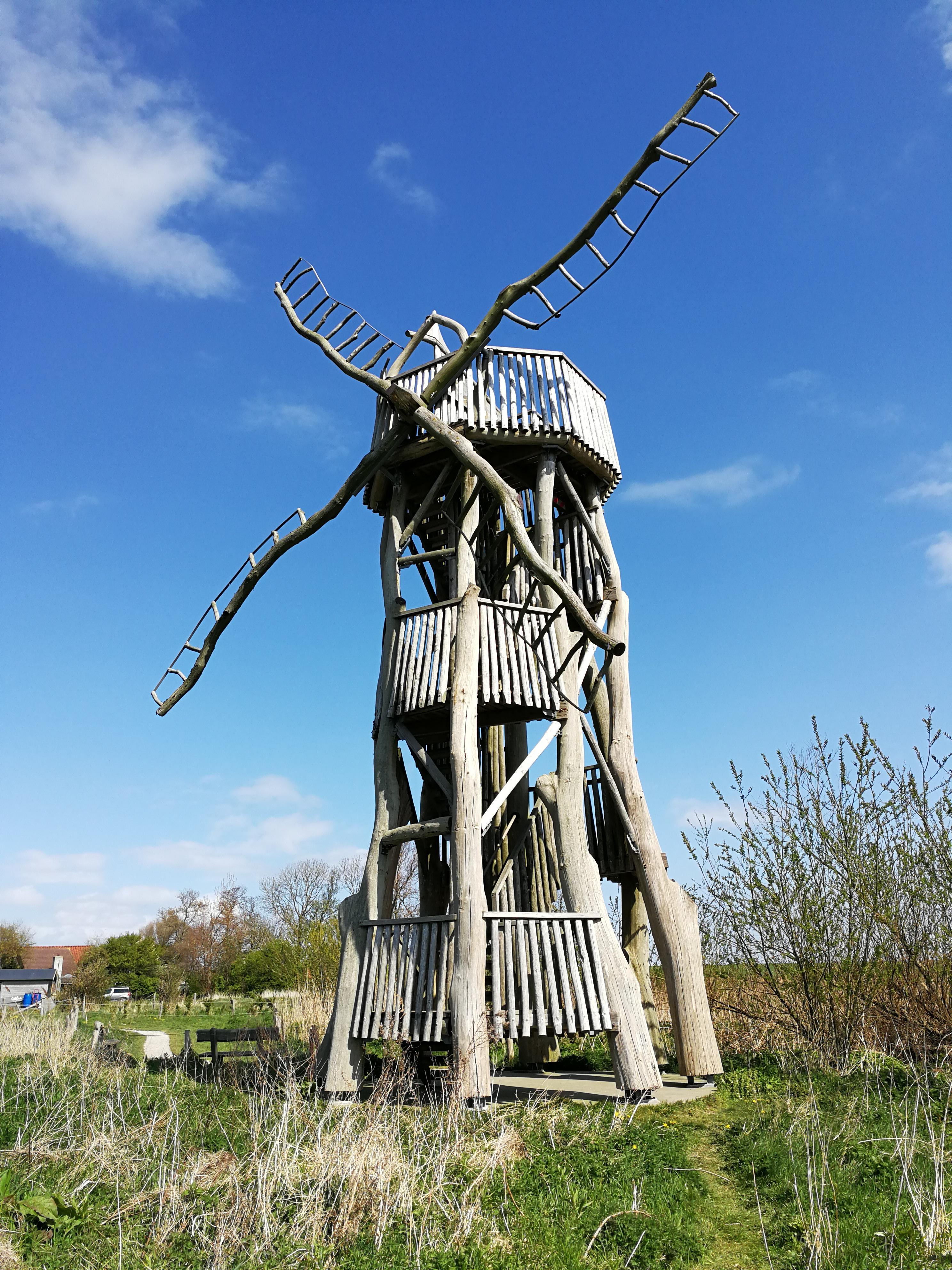 Wooden tower resembling a traditional windmill, featuring a multi-level structure with ladder-like railings, set under a blue sky with sparse clouds. The windmill has large, ladder-like vanes extending from the top. It is located in a grassy field with patches of untamed vegetation and sparse trees in the background.