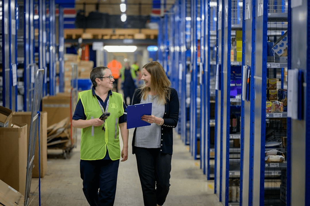 Two warehouse staff, one in a high-visibility vest, talking as they walk down a shelving aisle