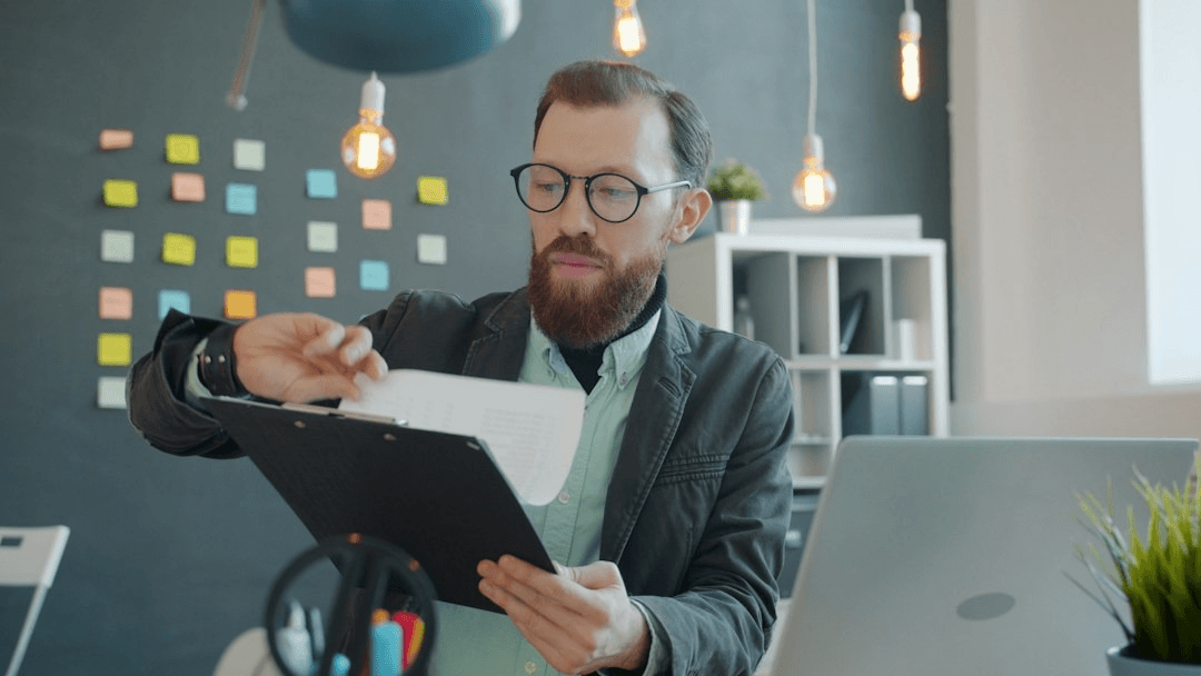 A man in glasses checking papers on a clipboard beside a laptop in an office