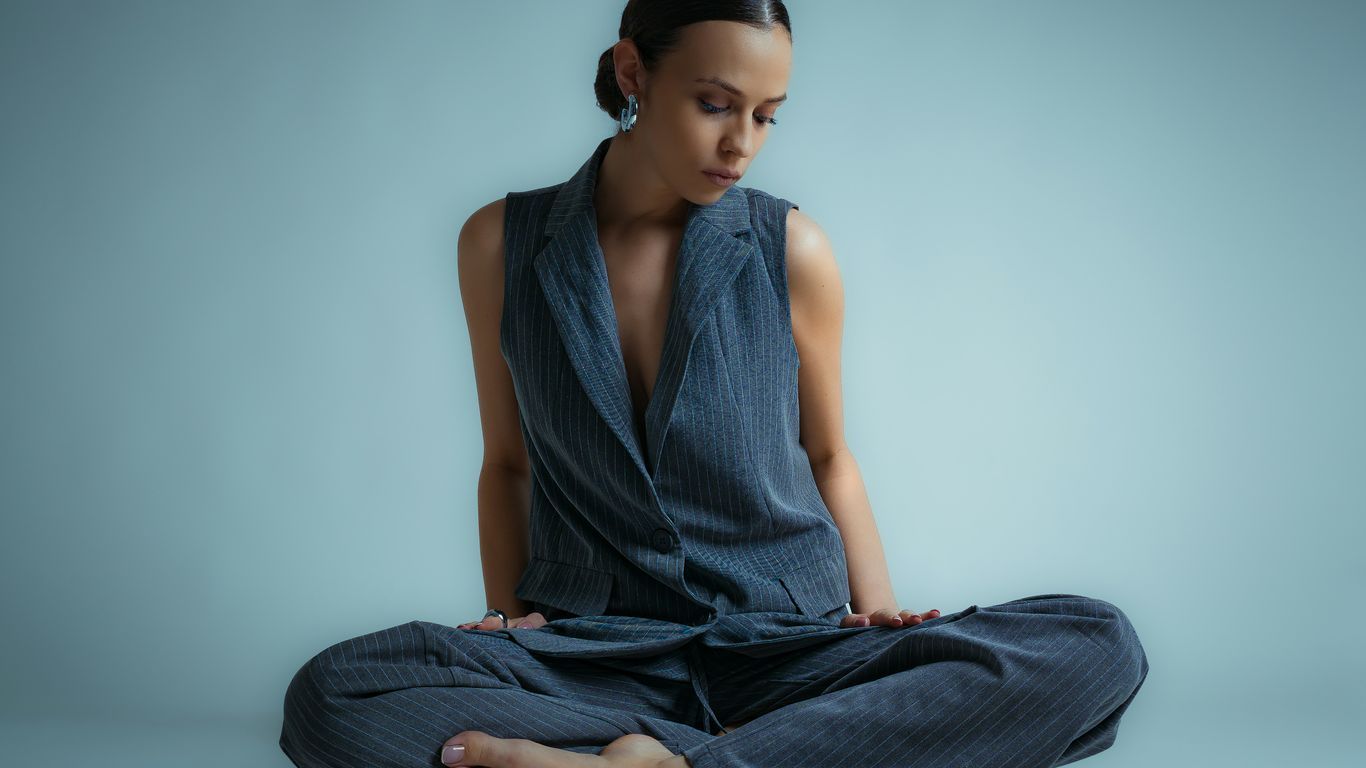A woman in a vest and pants sits cross-legged.