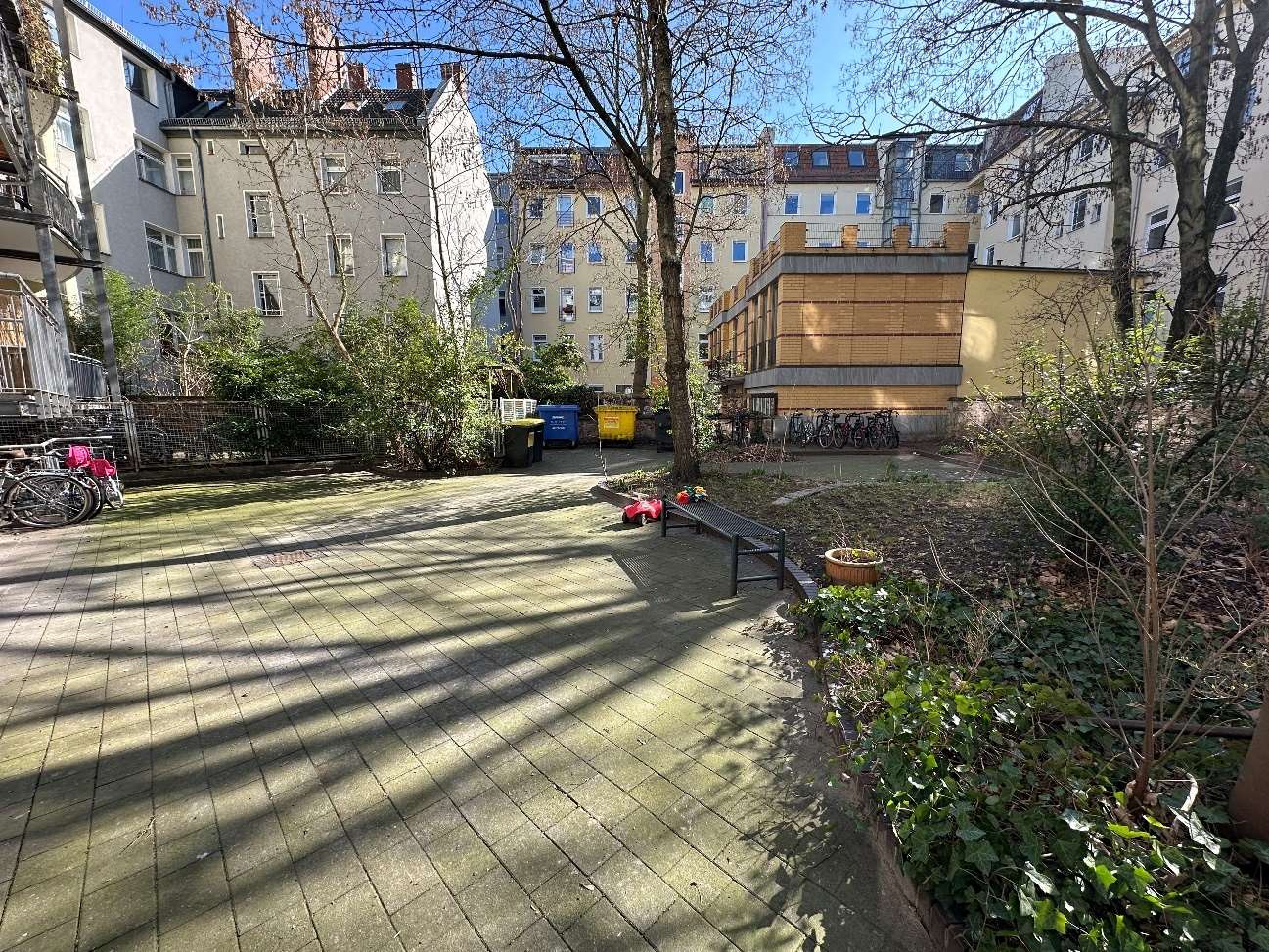 Gartenbereich mit Pflasterweg, Bäumen, Sträuchern und einer Bank vor mehrstöckigen Wohnhäusern in Berlin