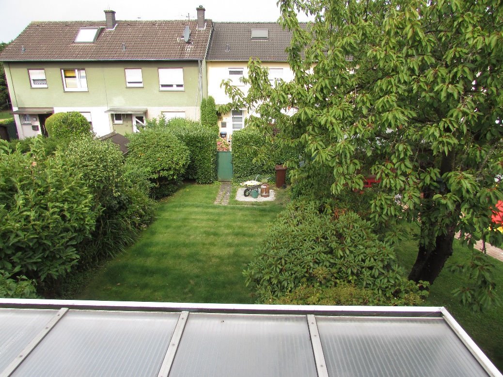 Grüner Garten mit Rasenfläche, Sträuchern und Bäumen, im Hintergrund Wohnhäuser mit braunen Dächern