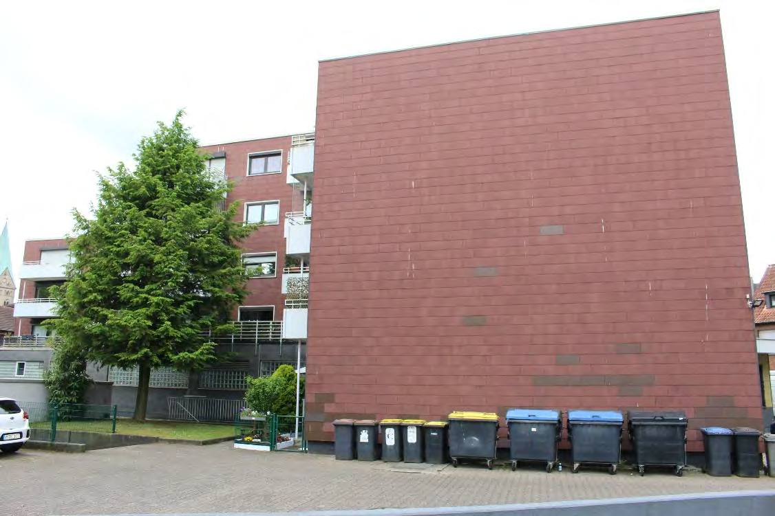 Rückansicht eines Mehrfamilienhauses mit rotem Backsteinmauerwerk, Mülltonnen im Vordergrund, Bäume und angrenzende Gebäude im Hintergrund