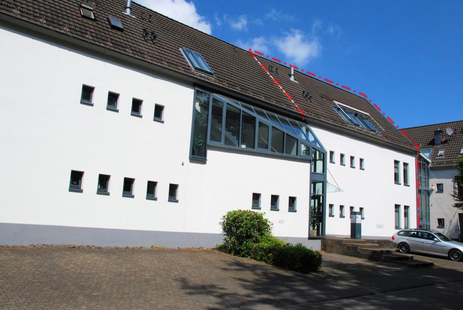 Seitenansicht eines weißen Gebäudes mit braunem Satteldach, Dachfenstern und moderner Glasverglasung, gepflastertem Vorplatz und geparktem Auto