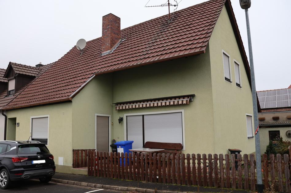 Seitenansicht eines Einfamilienhauses mit braunem Ziegeldach, hellgrüner Fassade, Fenstern mit Jalousien, Holzzaun und geparktem Auto vor dem Haus