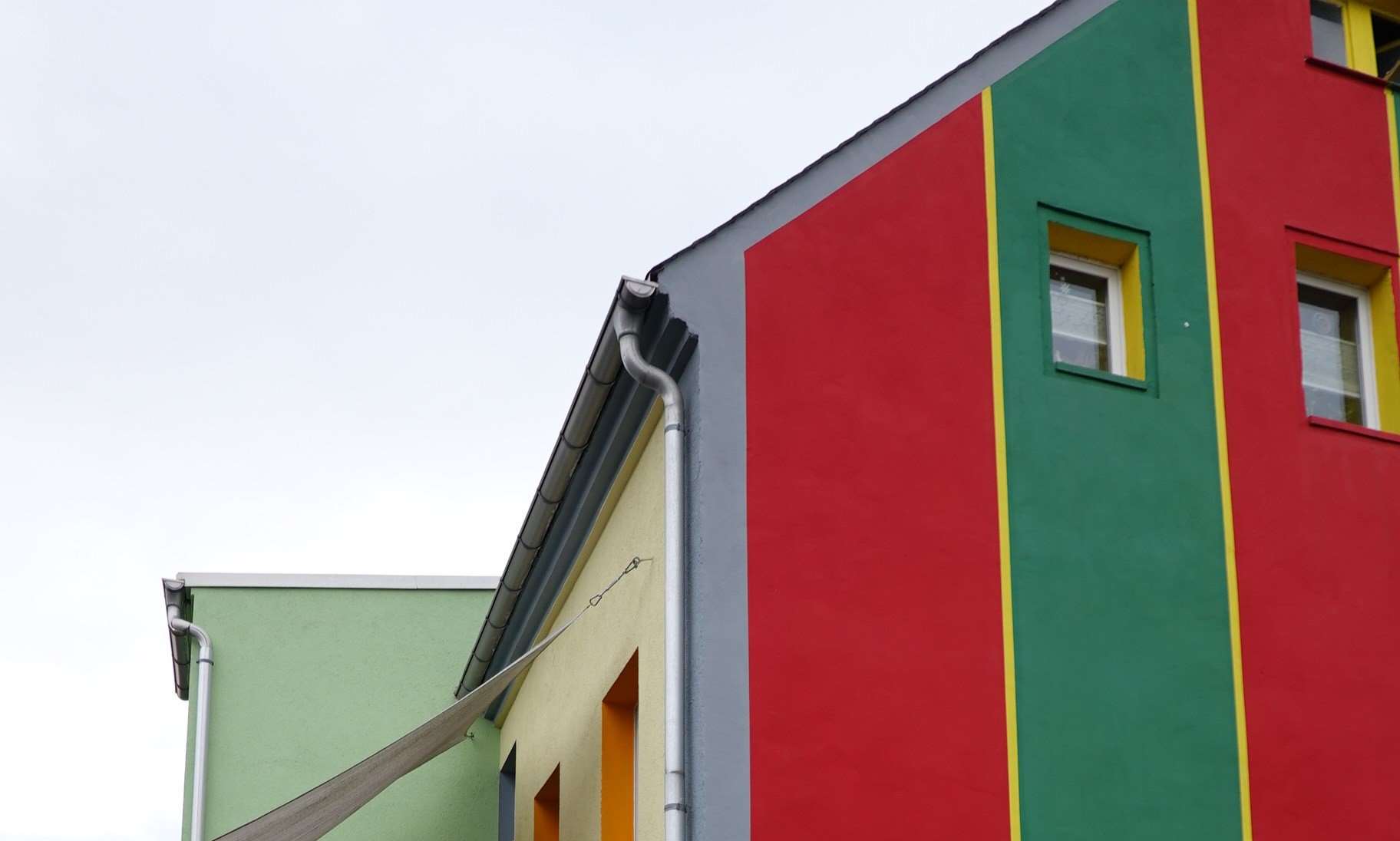 Seitenansicht eines farbenfrohen Gebäudes mit roten, grünen und gelben Fassadenelementen sowie Fenstern und Regenrohren