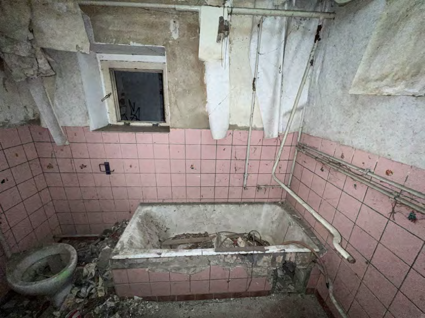 Verfallener Badezimmerbereich mit rosa Fliesen, zerstörter Badewanne, defekter Toilette und abblätternder Wandverkleidung. Fenster mit Gitter, sichtbare Rohrleitungen