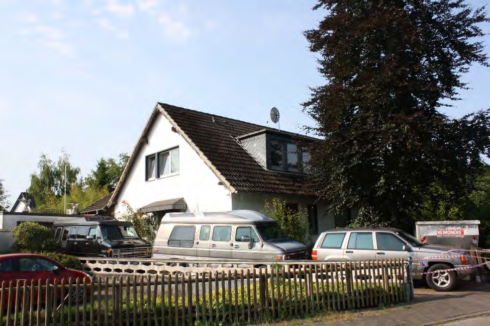 Zweifamilienhaus in Ratingen, An den Schlothen 9, 9a, mit dunklem Satteldach, weißen Wänden, Dachgaube und Satellitenschüssel; vorne geparkte Fahrzeuge und Holzzaun