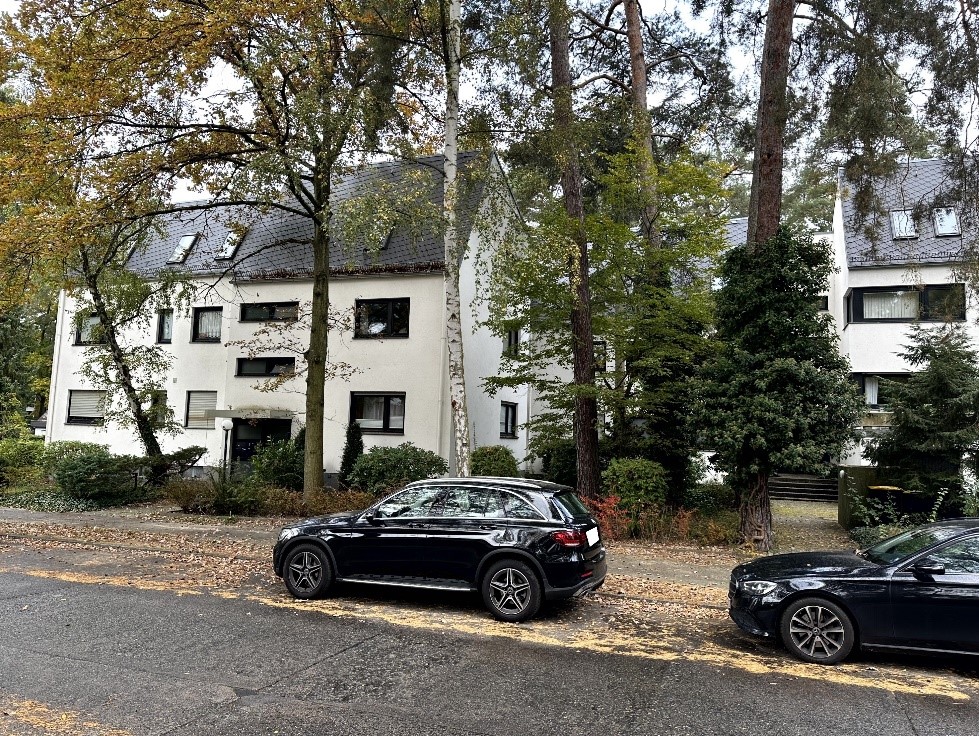 Seitenansicht eines weißen Mehrfamilienhauses mit dunklem Dach, umgeben von Bäumen und geparkten Autos an der Quermatenweg in Berlin