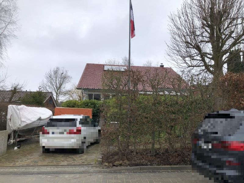 Rückansicht eines Einfamilienhauses in Hohwacht, Lütjenburger Straße 48, mit rotem Ziegeldach, Gartenhecke, geparktem Auto und Flaggenmast unter bewölktem Himmel
