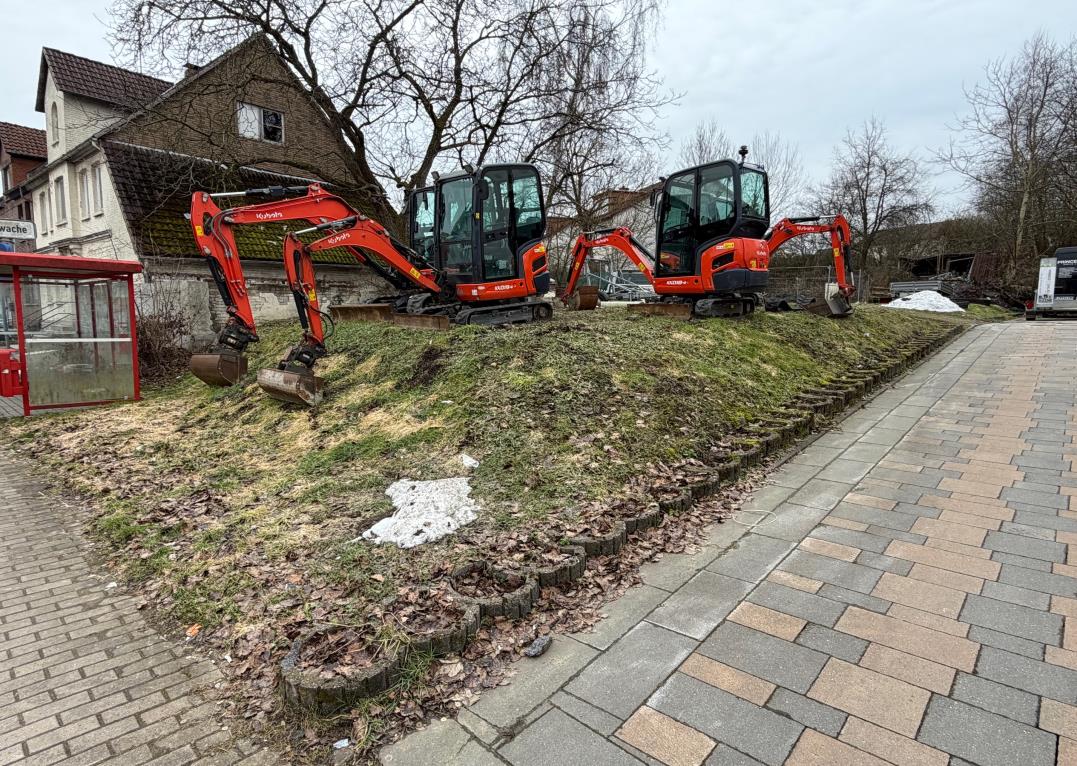 Gartenbereich mit mehreren roten Mini-Bagger auf Rasenfläche, gepflastertem Weg und Wohnhäusern im Hintergrund, Extertal, Bahnhofstraße 12
