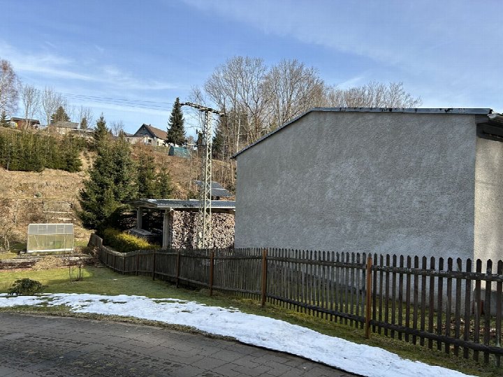 Gartenbereich mit Holzzaun, Schneeresten auf Rasen, Steinhaufen unter Dachüberstand und Blick auf bewaldeten Hang mit Wohnhäusern in Olbernhau, Rungstockstraße 120