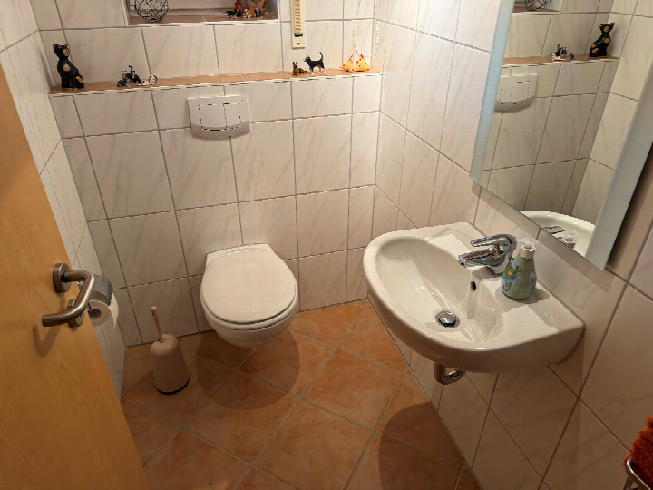Badezimmer mit weiß gefliesten Wänden, Wandwaschbecken, WC, Spiegel und braunen Fliesenboden. Dekoobjekte auf der Ablage über dem WC