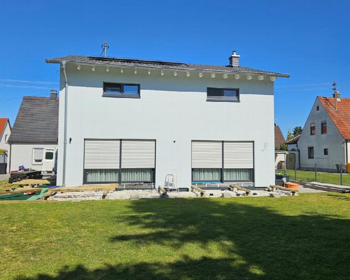Einfamilienhaus in Augsburg, Falkenweg 7a, mit weißer Fassade, dunklem Dach und Rollläden, vorne Grünfläche, im Hintergrund weitere Häuser