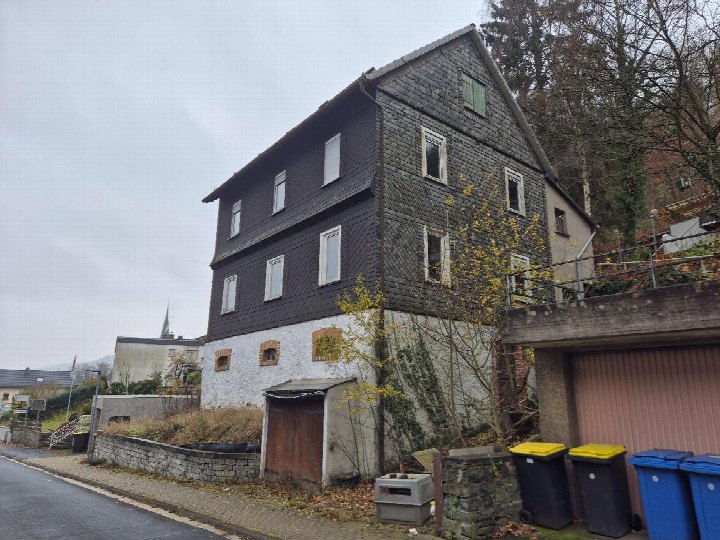 Seitenansicht eines Einfamilienhauses in Biedenkopf, Am Schloßhain, mit dunklem Schindeldach, weiß verputztem Erdgeschoss und Fenstern; im Vordergrund Mülltonnen und eine Mauer