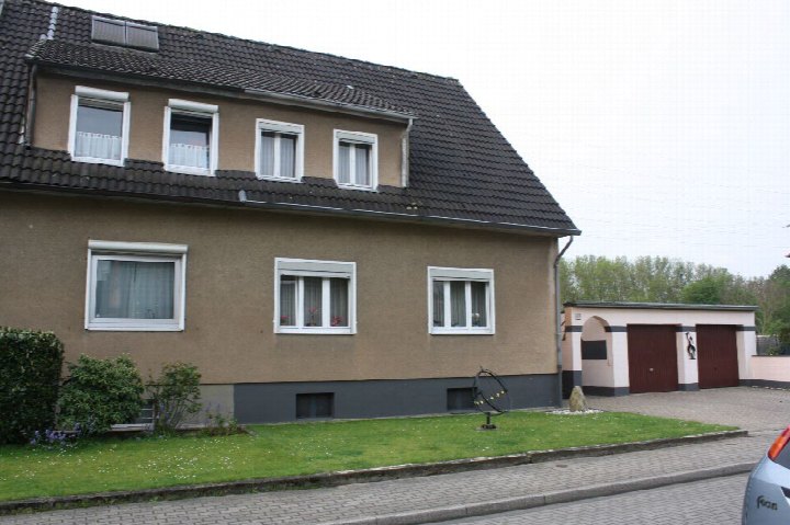 Zweifamilienhaus in Ratingen, Raiffeisenstraße 18, mit brauner Fassade, dunklem Satteldach und weißen Fenstern; Vorgarten mit Rasen und Hecke, rechts Nebengebäude mit Garagen