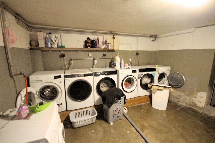 Keller mit mehreren Waschmaschinen, Wäschekörben und Regalen mit Reinigungsmitteln. Betonboden, sichtbare Rohrleitungen und niedrige Decke