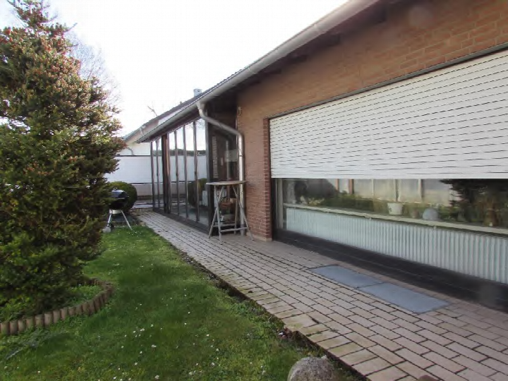 Garage mit weißer Rolltür, angrenzend an ein Backsteingebäude mit Glasfassade, gepflasterter Zugang und Grünfläche