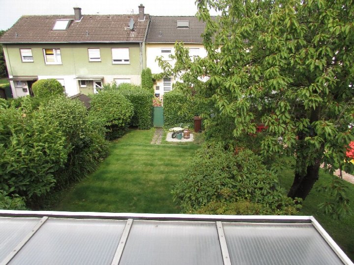 Grüner Garten mit Rasenfläche, Sträuchern und Bäumen, im Hintergrund Wohnhäuser mit braunen Dächern