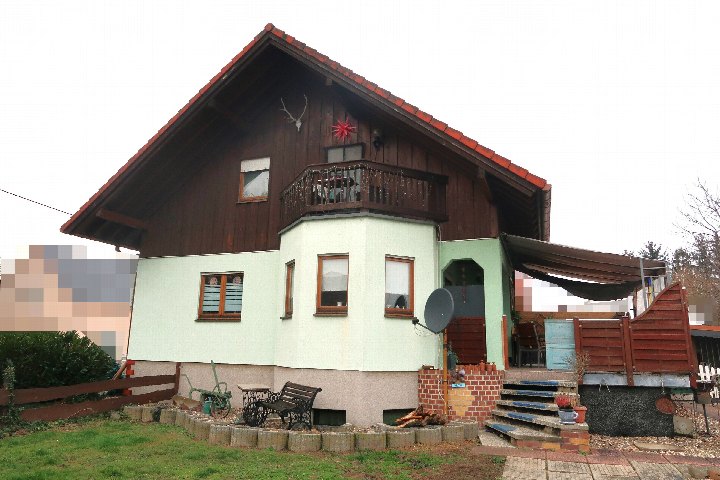 Einfamilienhaus mit dunklem Holzobergeschoss, hellgrüner Fassade, Balkon und Vorgarten. Eingangsbereich mit Treppe und Sitzgelegenheit