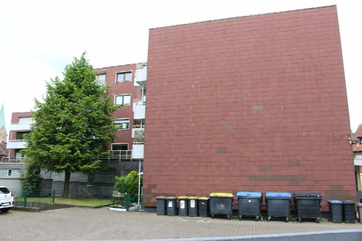 Rückansicht eines Mehrfamilienhauses mit rotem Backsteinmauerwerk, Mülltonnen im Vordergrund, Bäume und angrenzende Gebäude im Hintergrund