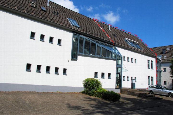 Seitenansicht eines weißen Gebäudes mit braunem Satteldach, Dachfenstern und moderner Glasverglasung, gepflastertem Vorplatz und geparktem Auto