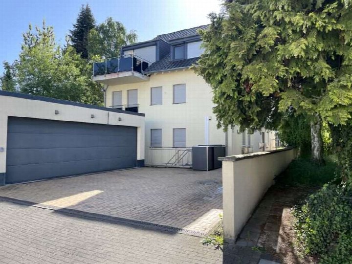 Gebäude mit Anbau und Garagenanlage, gepflasterter Vorplatz, Sicht auf Wohnhaus mit Balkon und Bäumen im Hintergrund