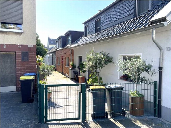 Hinterhofansicht eines Einfamilienhauses mit grünem Gittertor, Mülltonnen und Pflanzen in Kübeln, umgeben von Wohnhäusern in Köln-Niehl