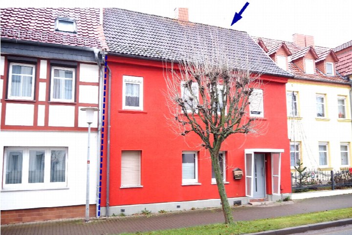 Vorderansicht eines roten Einfamilienhauses mit Satteldach, Fenstern und Eingang, umgeben von Nachbarhäusern und einem Baum vorne