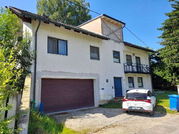 Zweistöckiges Wohnhaus mit brauner Garagentür, blauer Eingangstür und Balkon im Obergeschoss, geparktes weißes Auto vor dem Haus